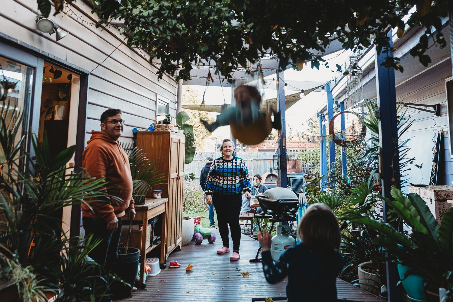 Family enjoying time on a plant-filled outdoor patio with a child swinging in the background.