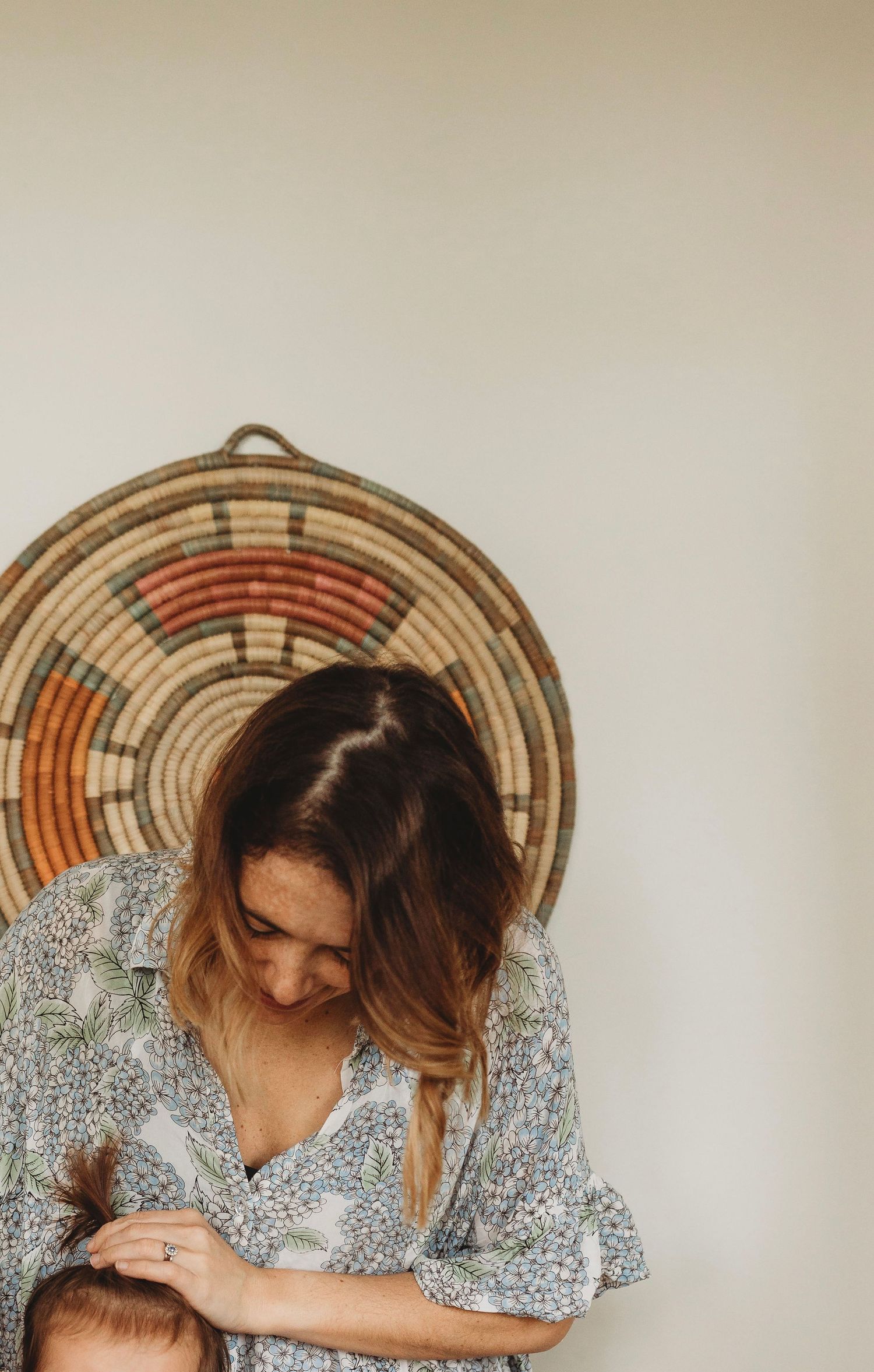 Woman in floral dress looking down while holding a baby with woven basket wall art behind her.