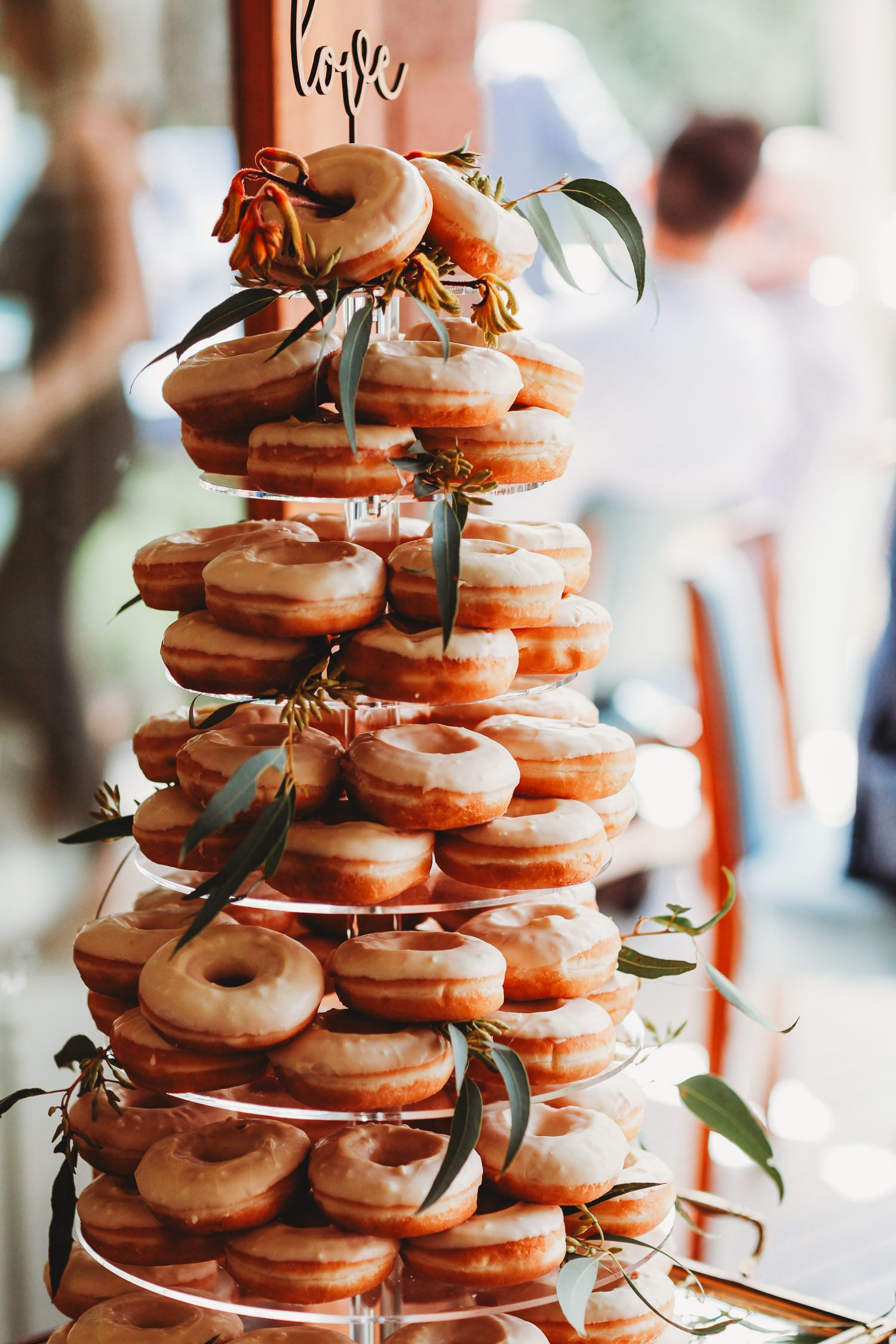 A towering wedding donut cake decorated with greenery and flowers at a reception.