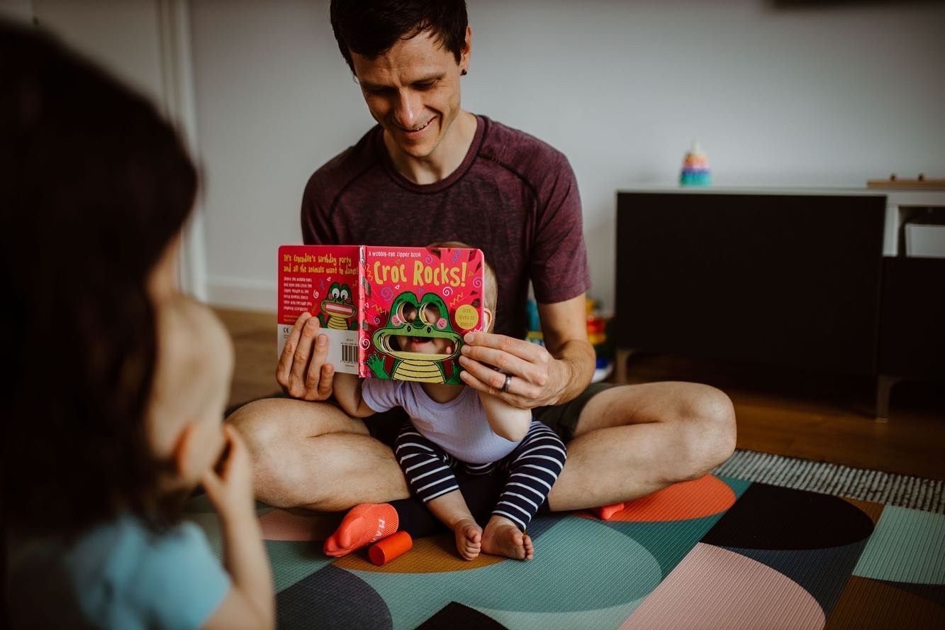 Smiling father reading a colorful childrens book to toddler while sitting cross-legged on yoga mat.