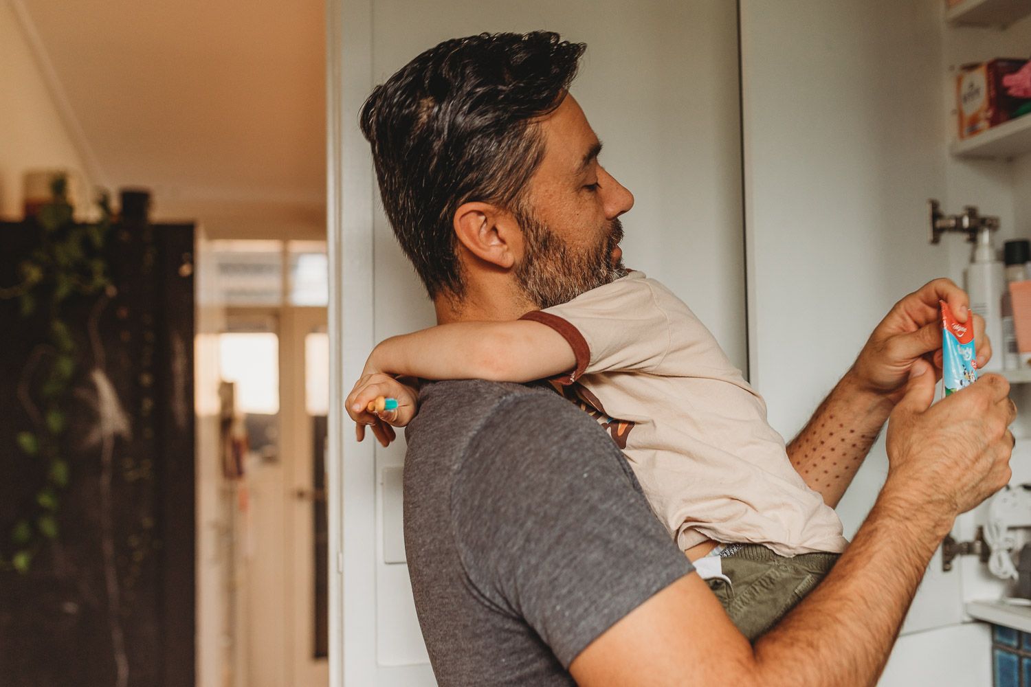 A bearded father holds his baby while reaching into a cabinet in a bright home interior.