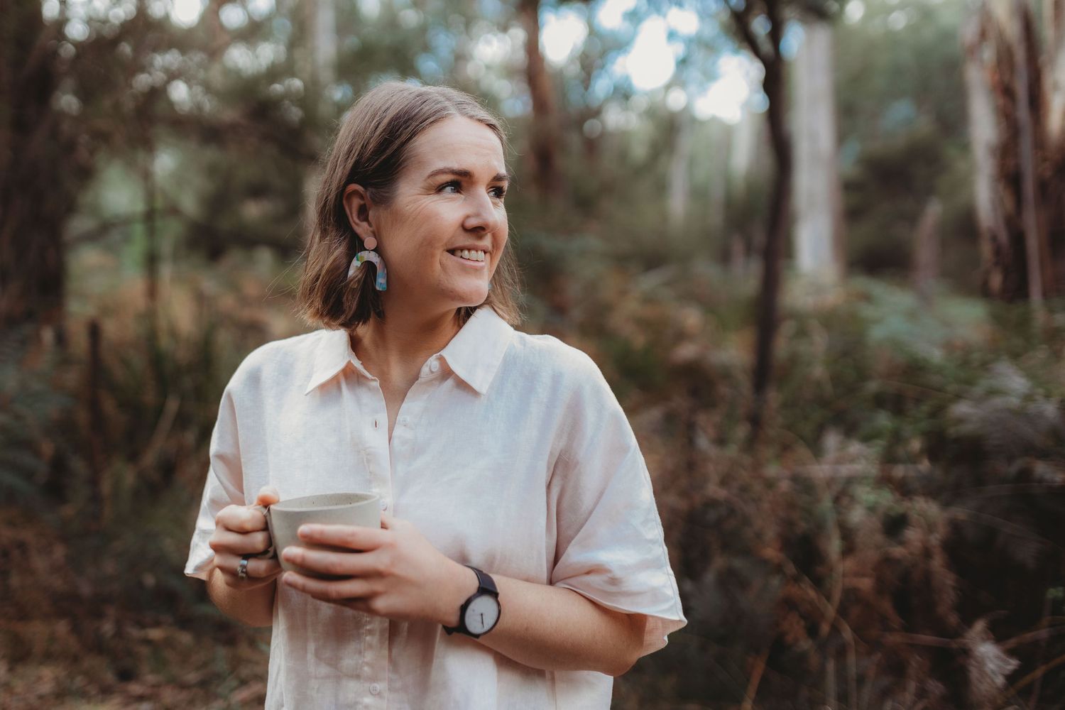 Smiling woman holding a reusable cup outdoors in a lush green forest setting.