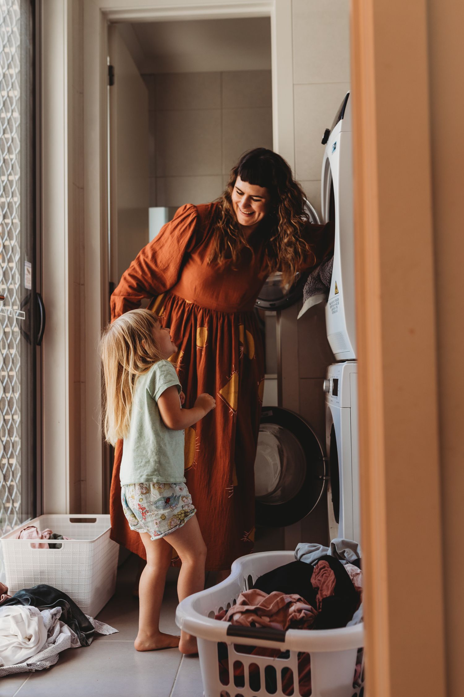 Mother in rust dress smiles at toddler daughter while doing laundry together near washing machine.