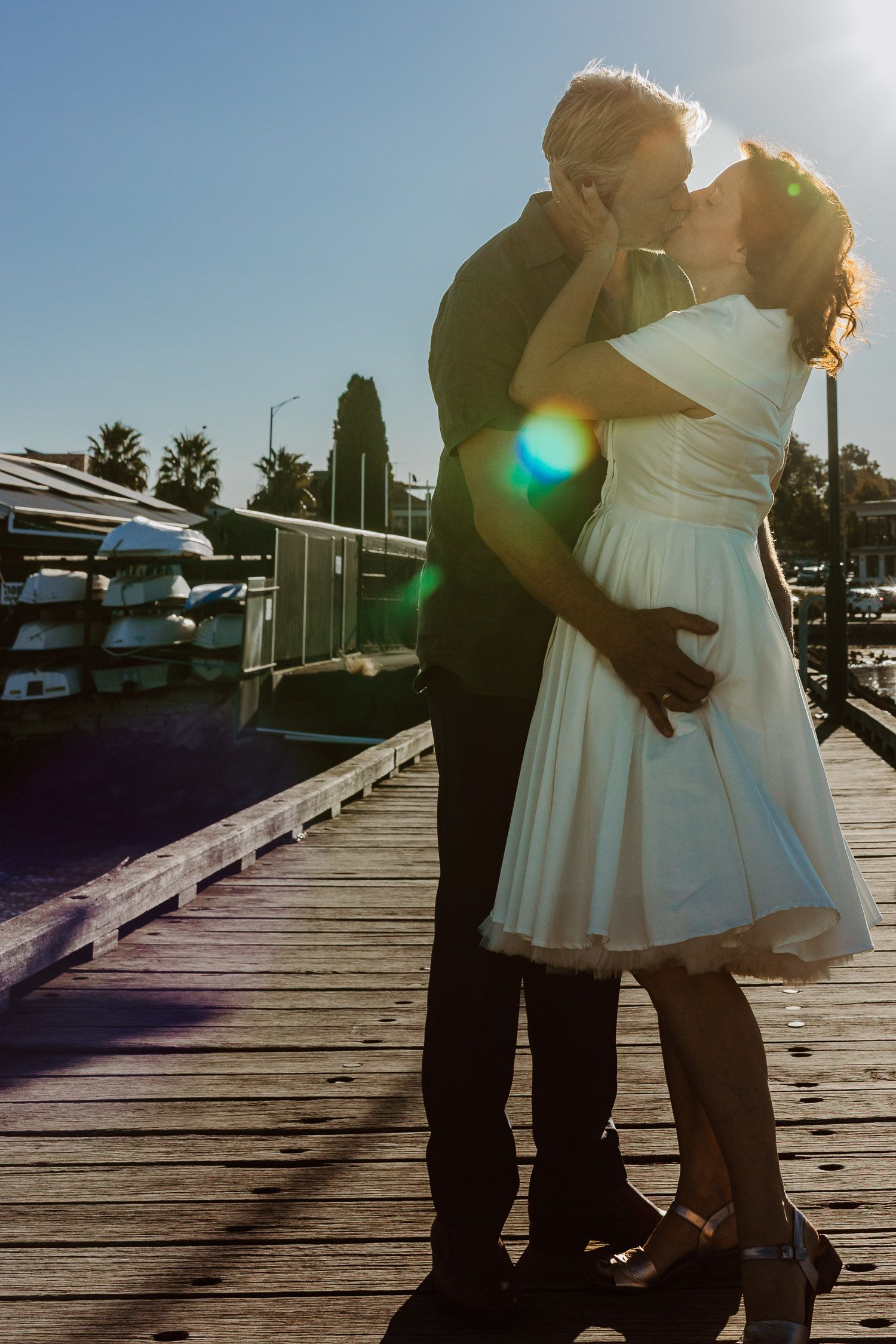 Bride and groom sharing a romantic kiss on a sunny marina dock with lens flare in a wedding photo.