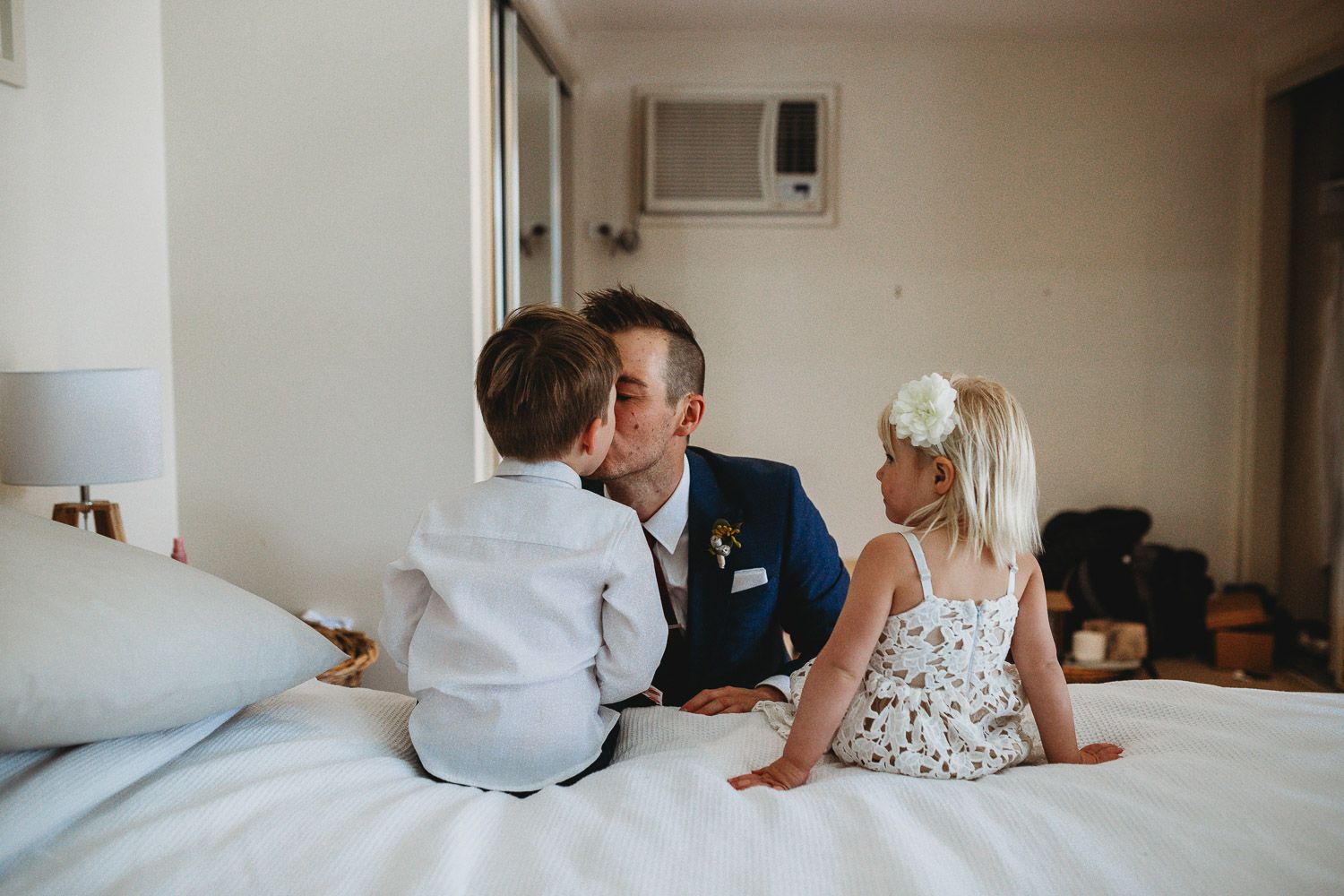 Groom in blue suit sharing tender moment with two children on hotel bed.