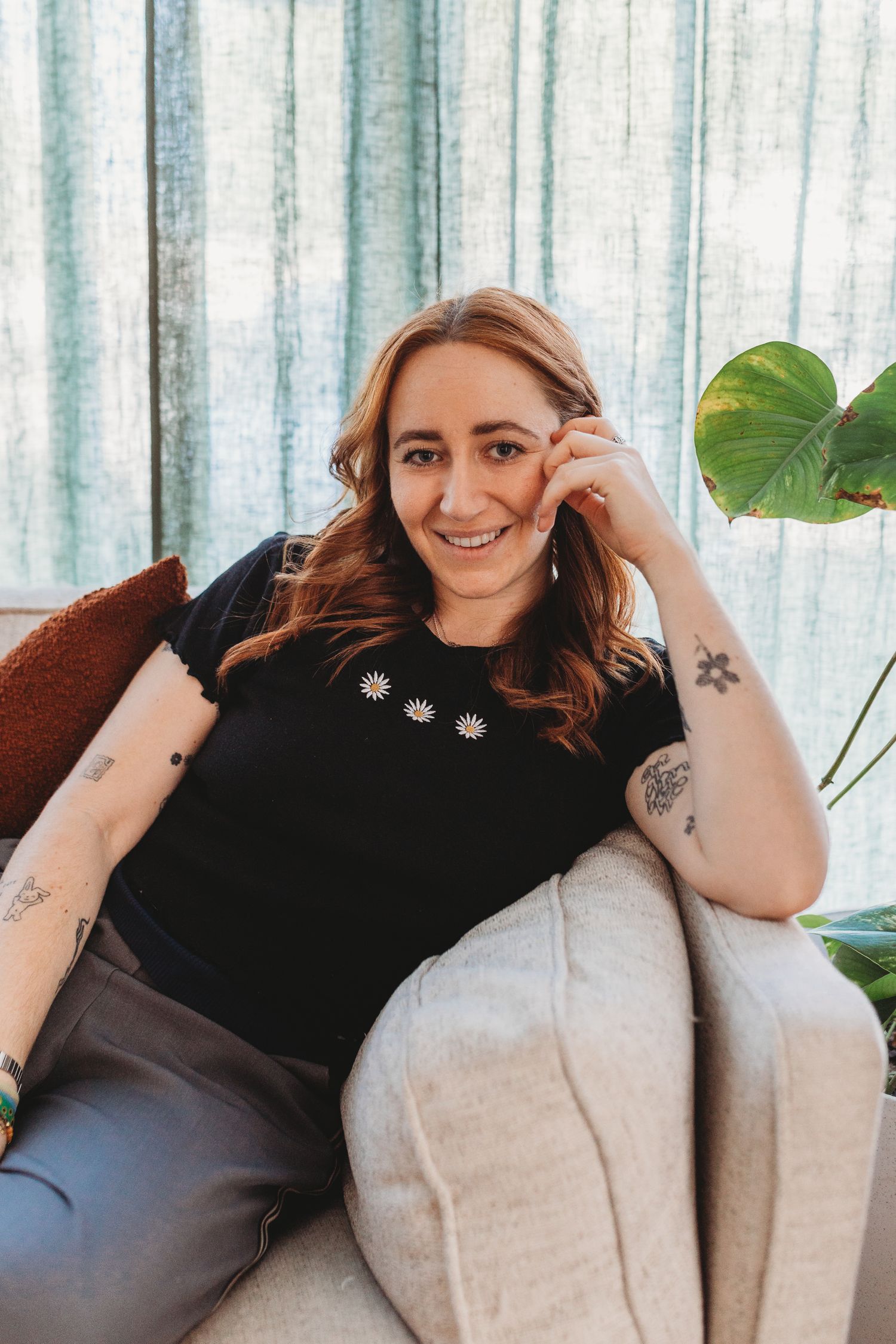 Smiling woman with tattoos relaxing in a light grey armchair beside a monstera plant with teal curtains behind.