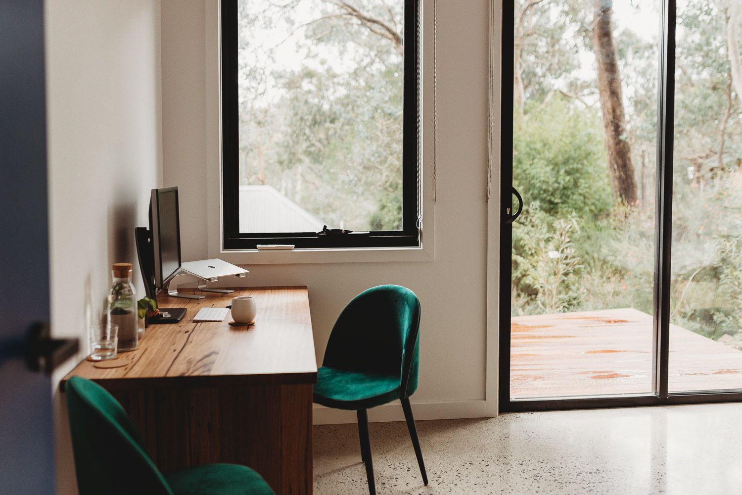 Modern home office with wooden desk, green velvet chairs, and large windows overlooking lush green trees.