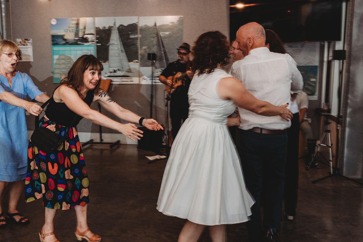Wedding guests dancing and celebrating with bride in white dress at lively indoor reception.