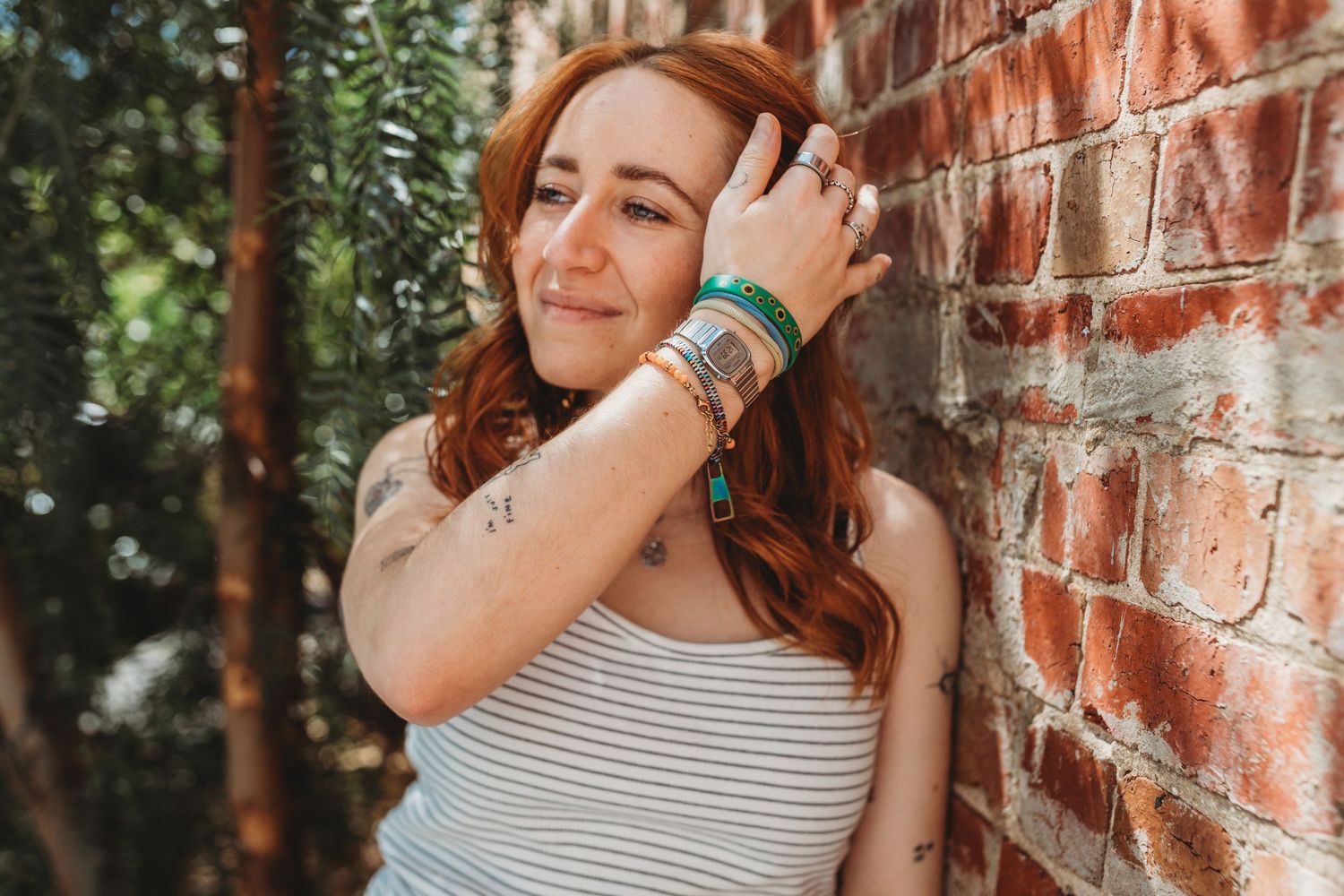 Smiling redhead woman with tattoos and bracelets leaning against brick wall outdoors.