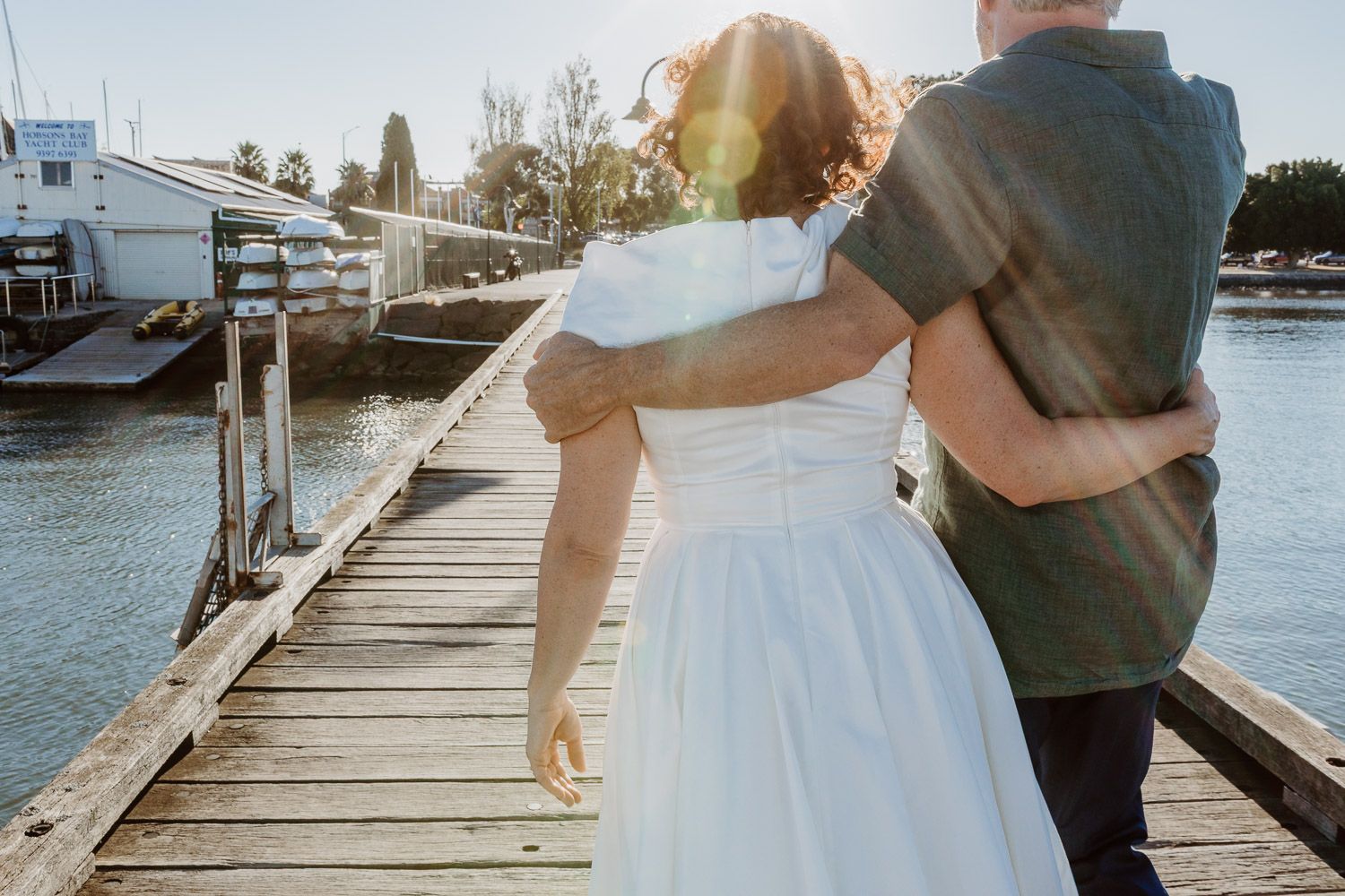 Bride and groom walking arm in arm along a sunny marina boardwalk with sailboats in the background.