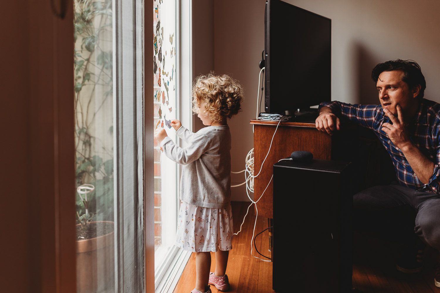 A father sits near a TV while his young daughter looks out a large window at home.