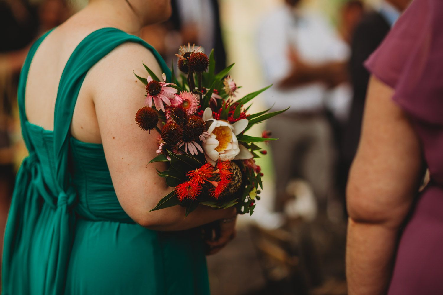 Bridesmaid in teal green dress holding a colorful wildflower bouquet at an outdoor wedding.