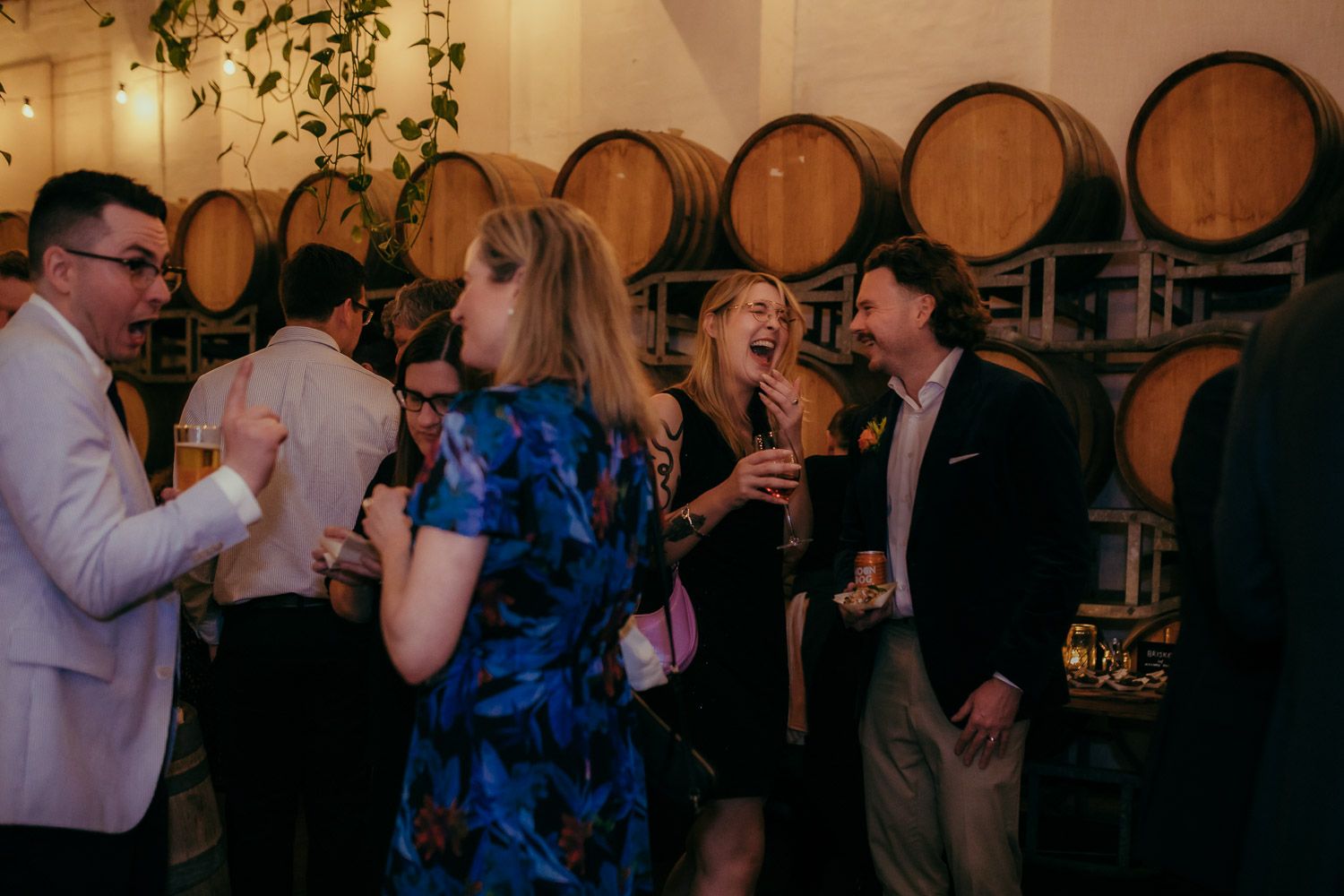 Guests laughing and socializing in a rustic venue surrounded by oak wine barrels.