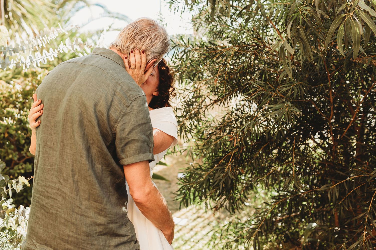 Older couple sharing an intimate embrace outdoors surrounded by green foliage in warm sunlight.