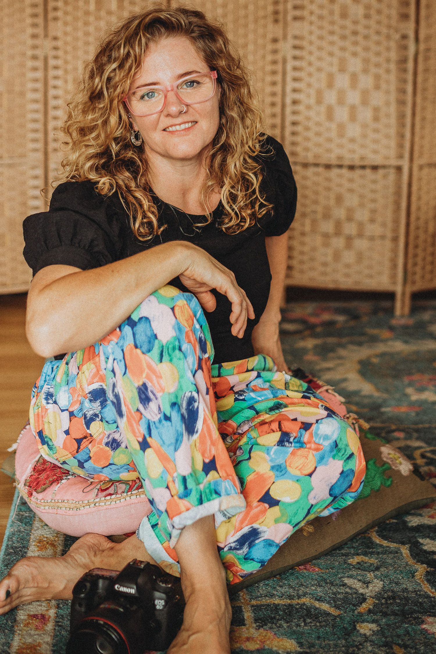 Woman with curly hair and pink glasses sits cross-legged on a rug wearing colorful pants, with a Canon camera nearby.