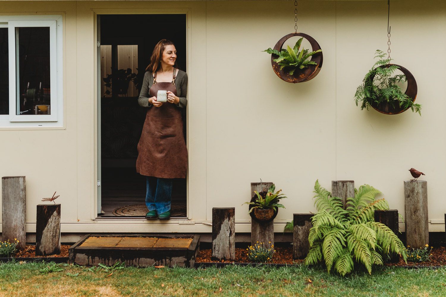 Smiling woman in leather apron holding coffee mug standing in doorway of rustic home with plants.