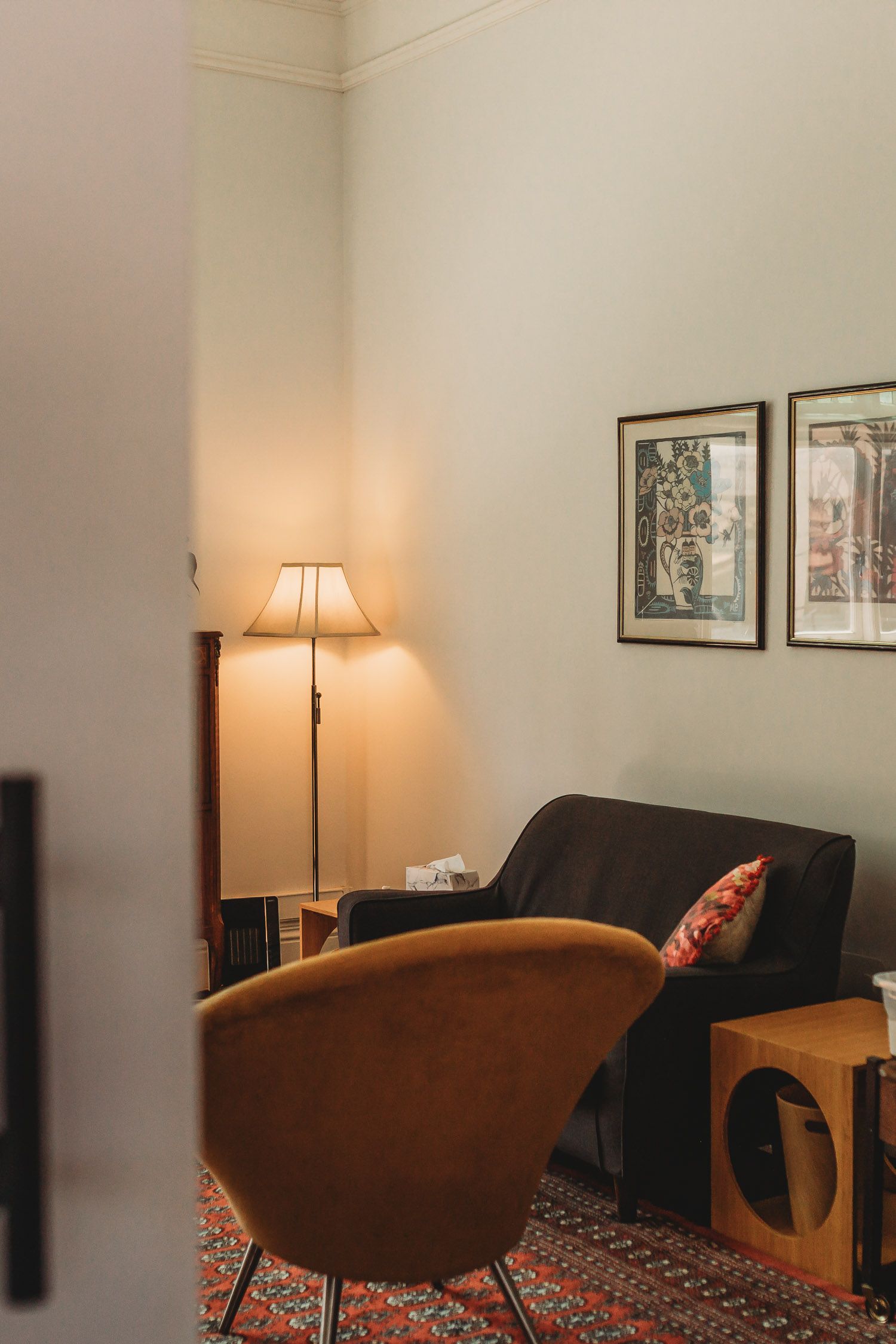 Cozy therapy room with warm floor lamp, dark sofa, orange chair, and framed artwork on walls.