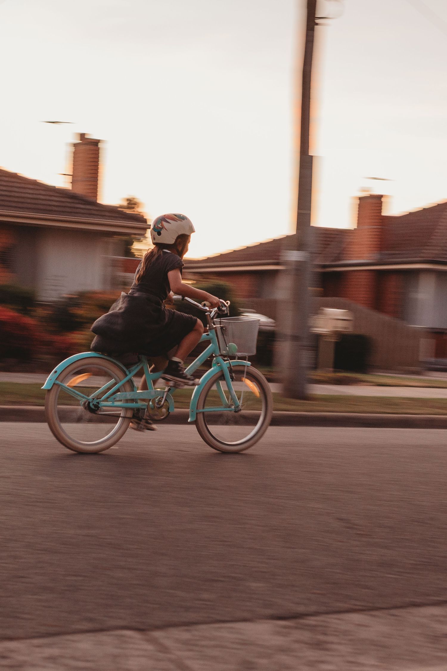 Child riding a teal bicycle wearing a white helmet through a suburban street at sunset.