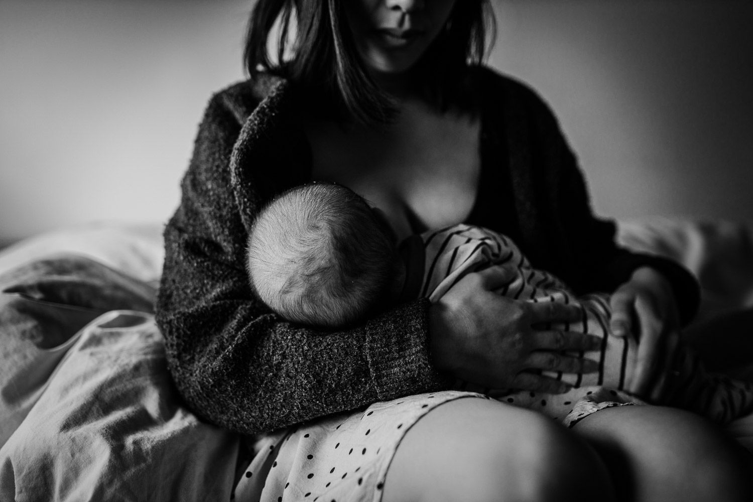 Black and white photo of mother breastfeeding newborn baby while sitting on bed.