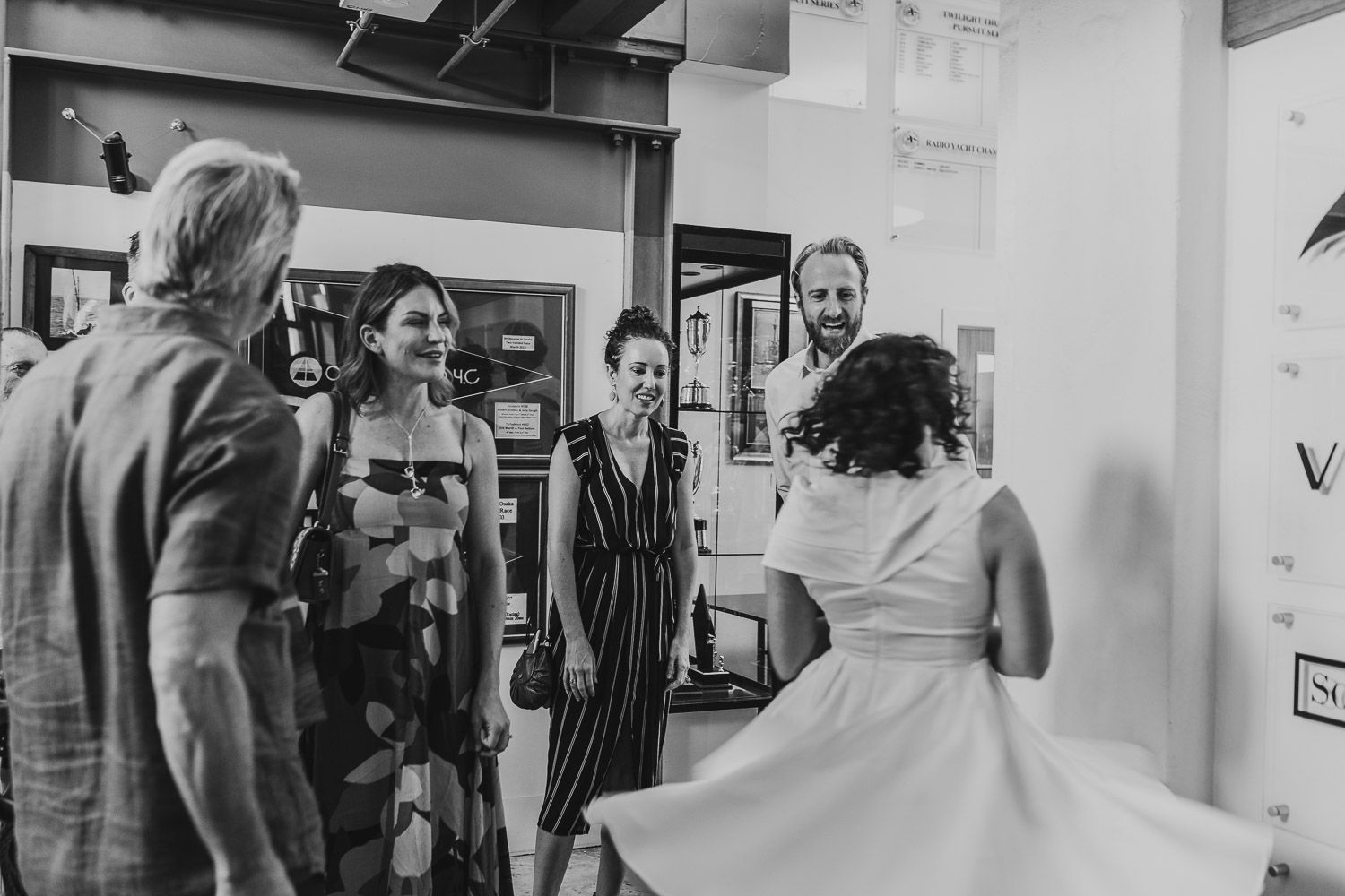 Black and white photo of a bride in a full skirt twirling while greeting smiling guests at her wedding reception.