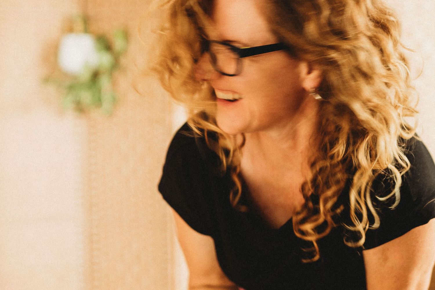Woman with curly blonde hair and glasses smiling softly in warm natural light.