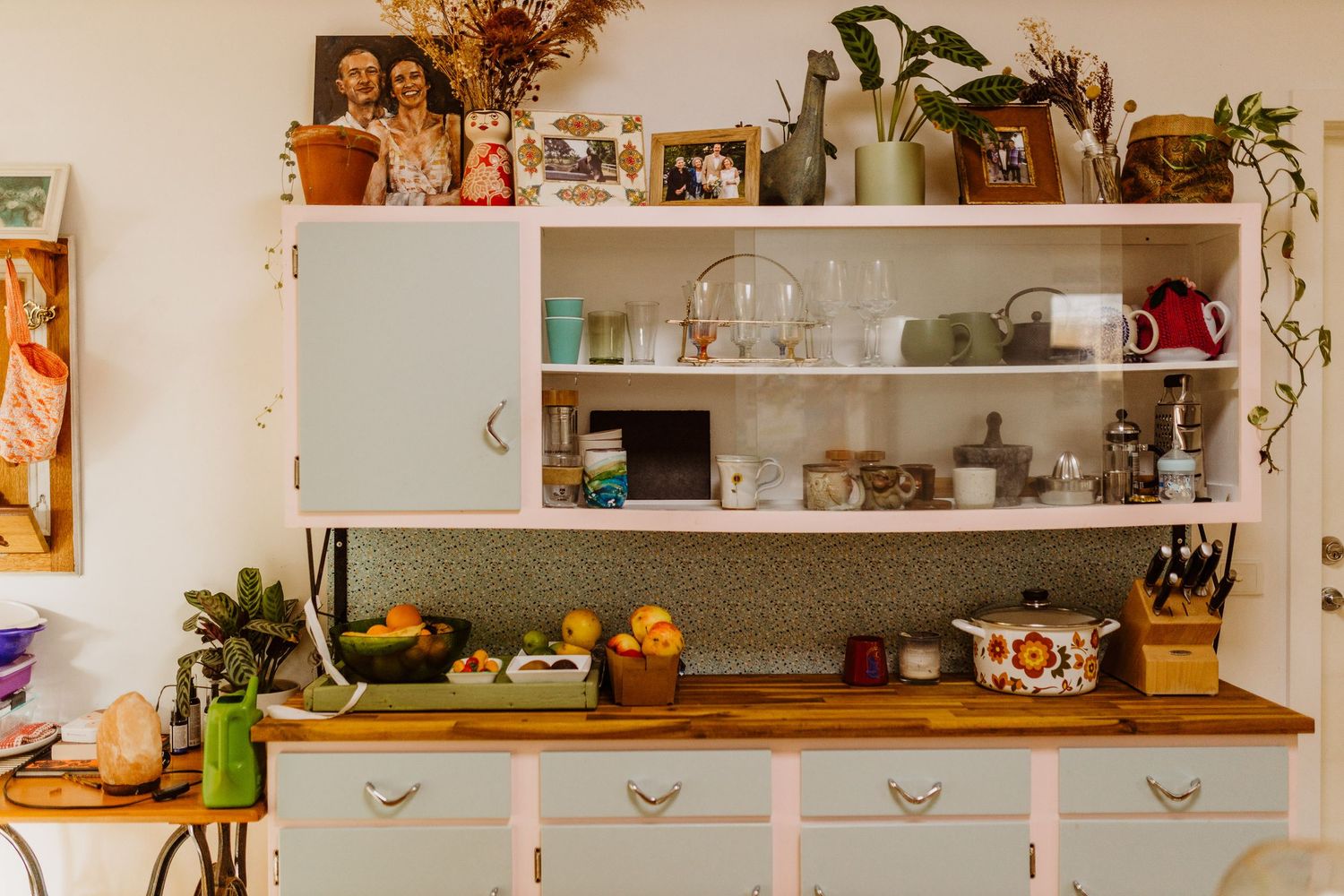 Cozy kitchen corner with a wooden countertop, white open shelves holding dishes and plants, a red teapot, fresh fruit and family photos.