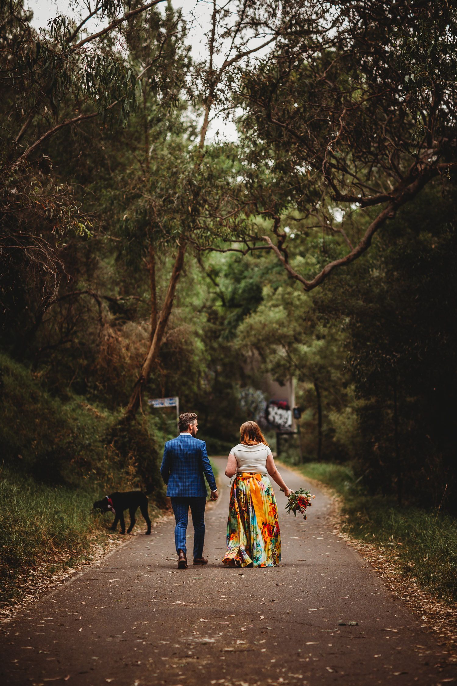 Bride in colorful floral skirt and groom in blue suit walking away on a forest path.