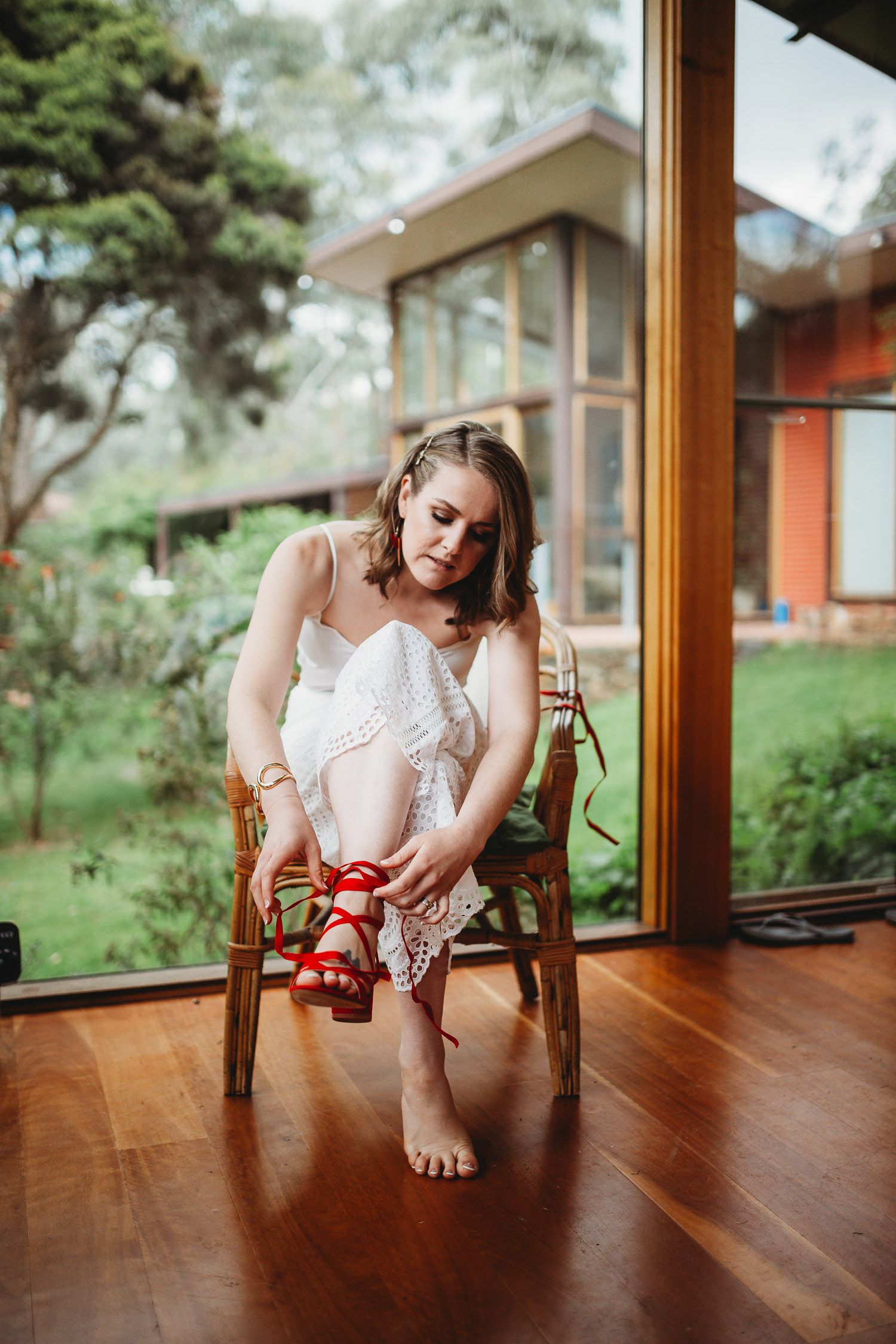 Bride in white lace dress putting on red heeled sandals while seated.