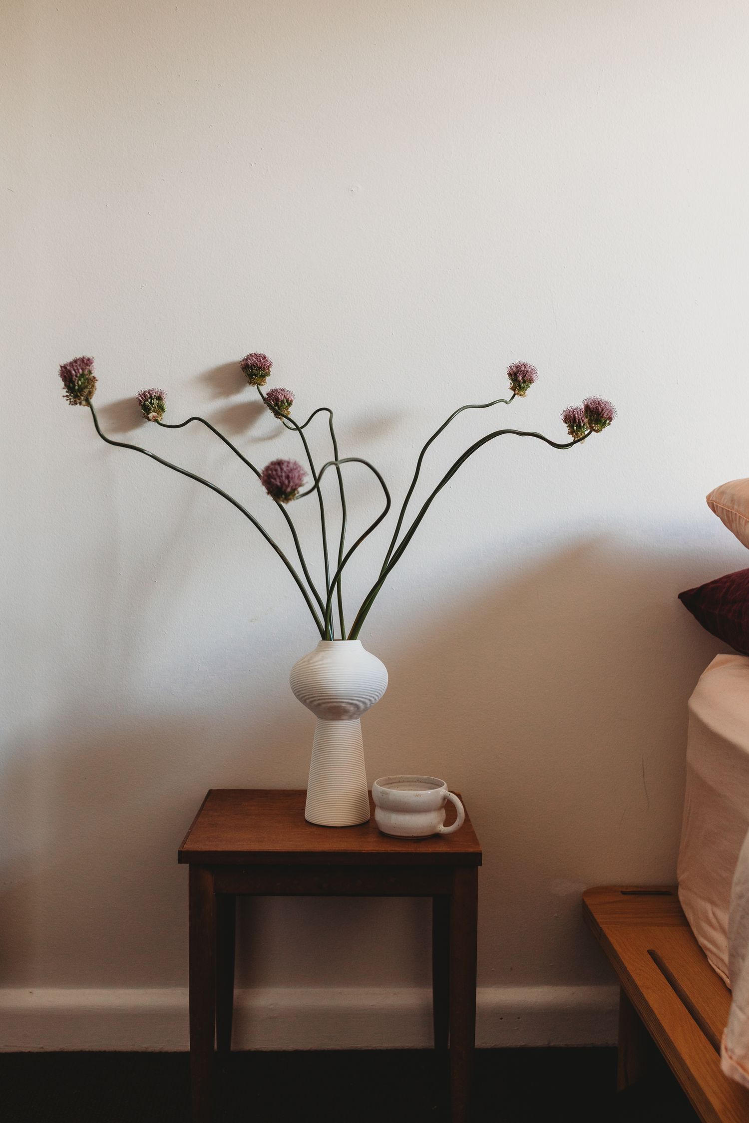 White ceramic vase with allium flowers on a wooden side table beside a mug and bed.