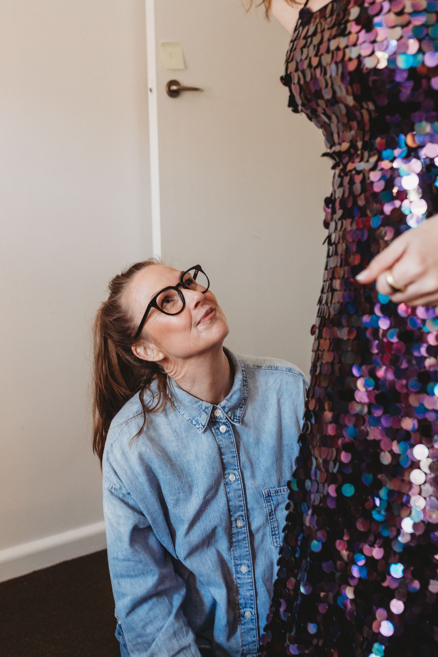 Fashion designer in glasses kneeling to inspect sequin dress hem detail closely.
