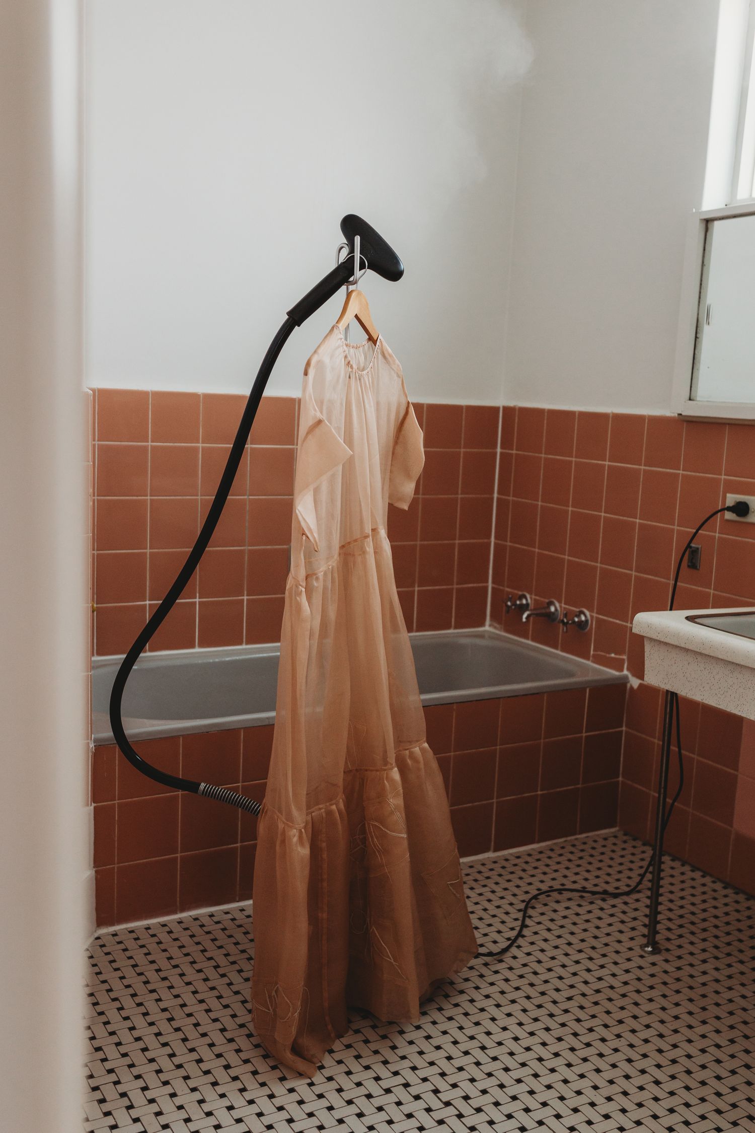 A peach tiered dress hanging from a steamer in a vintage bathroom with terracotta tiles.