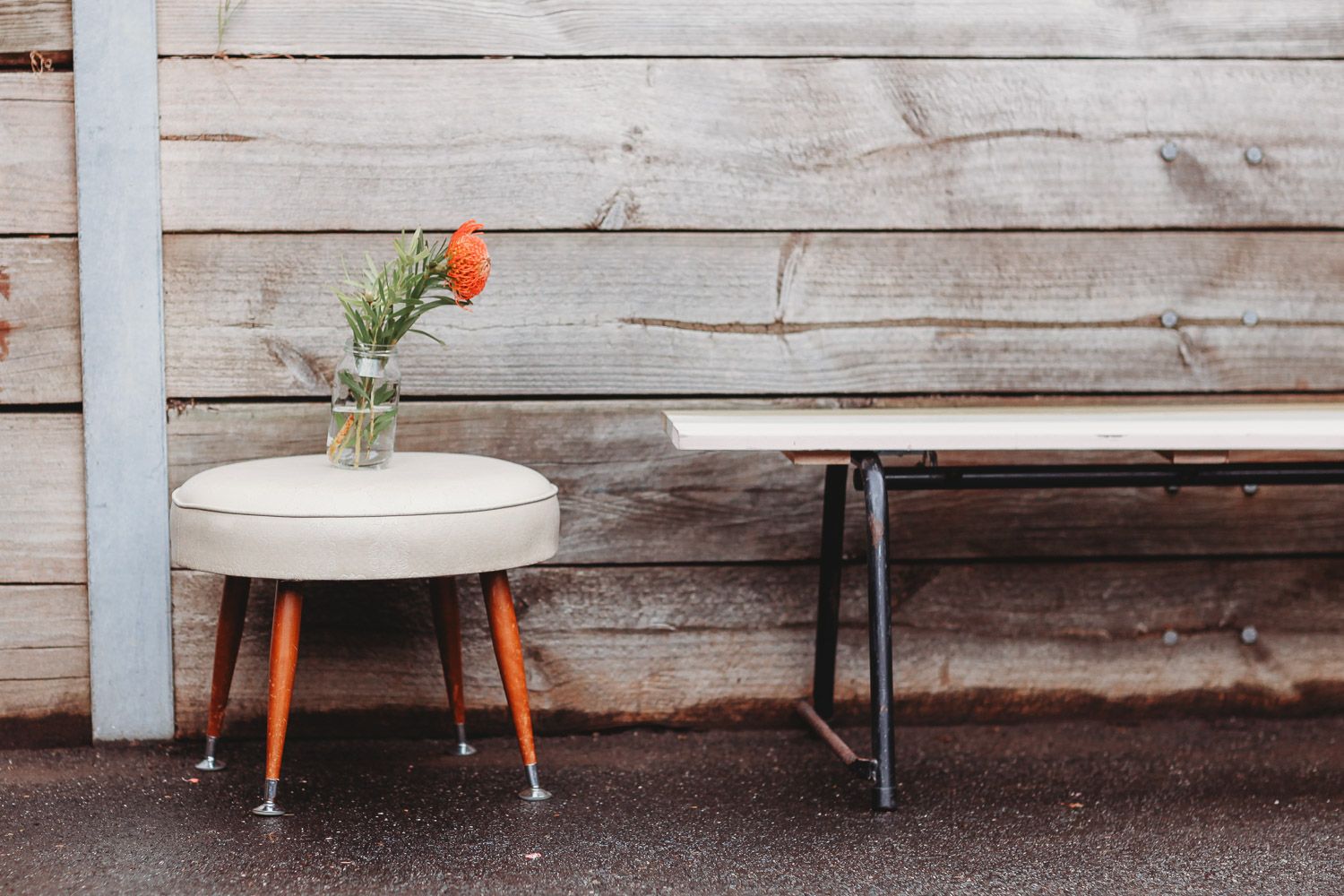 Vintage stool with orange flower vase beside rustic wooden bench outdoors.