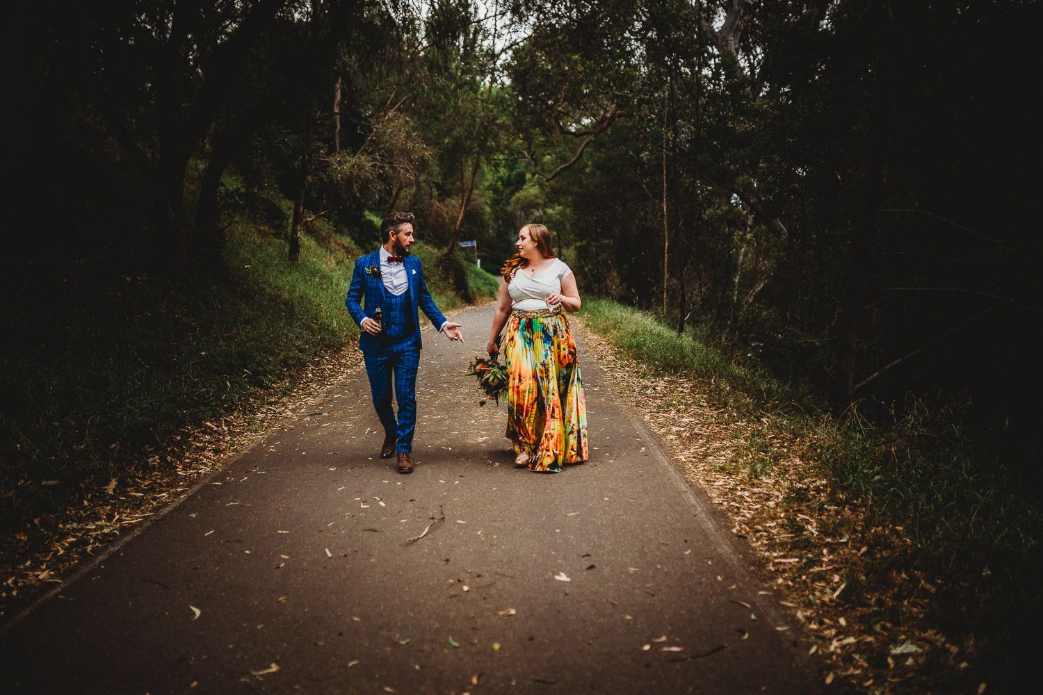 Bride in vibrant floral skirt and groom in blue suit strolling hand in hand on a forest path.