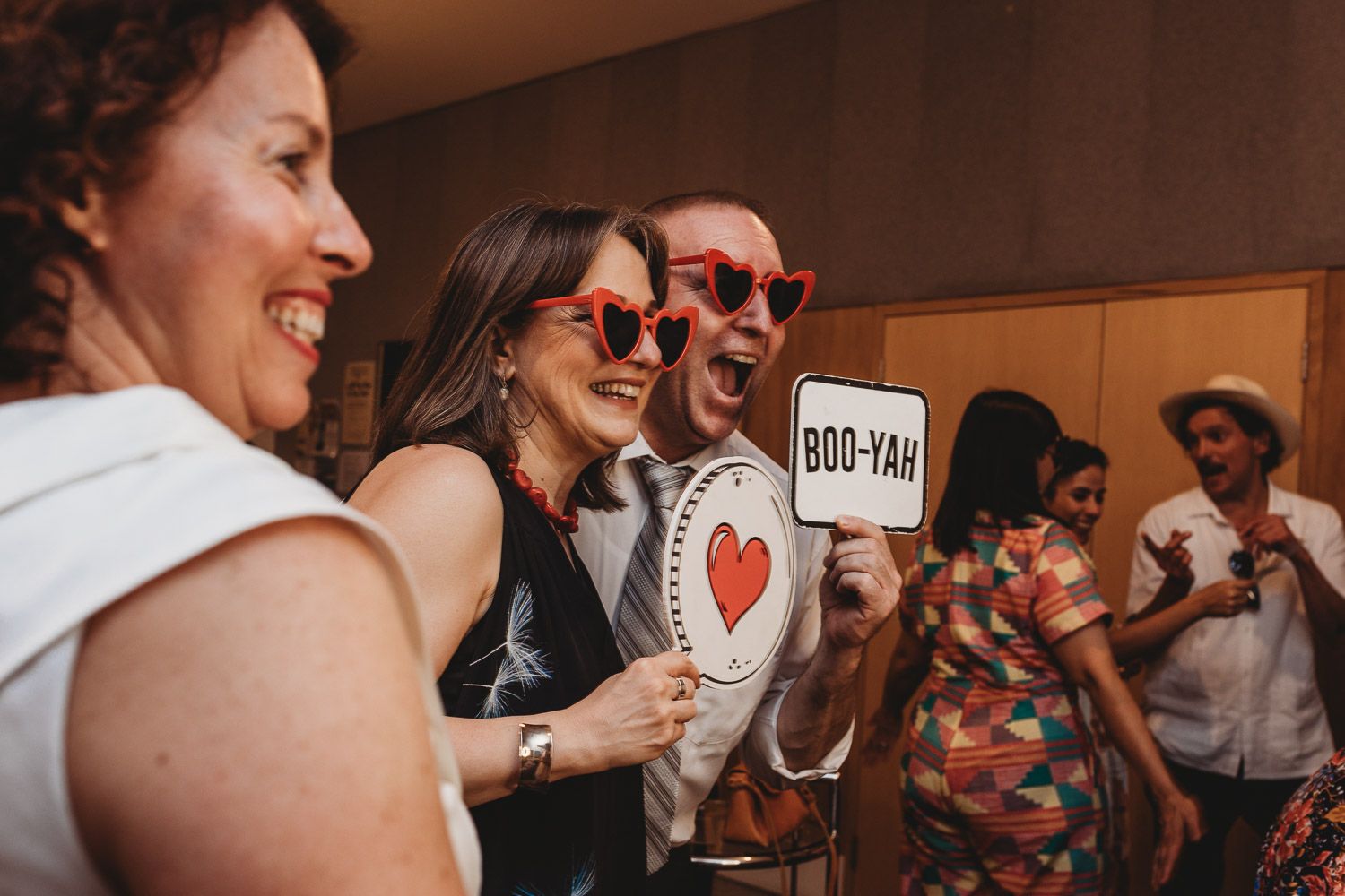 Wedding guests laughing and posing with heart-shaped props and fun signs at reception.