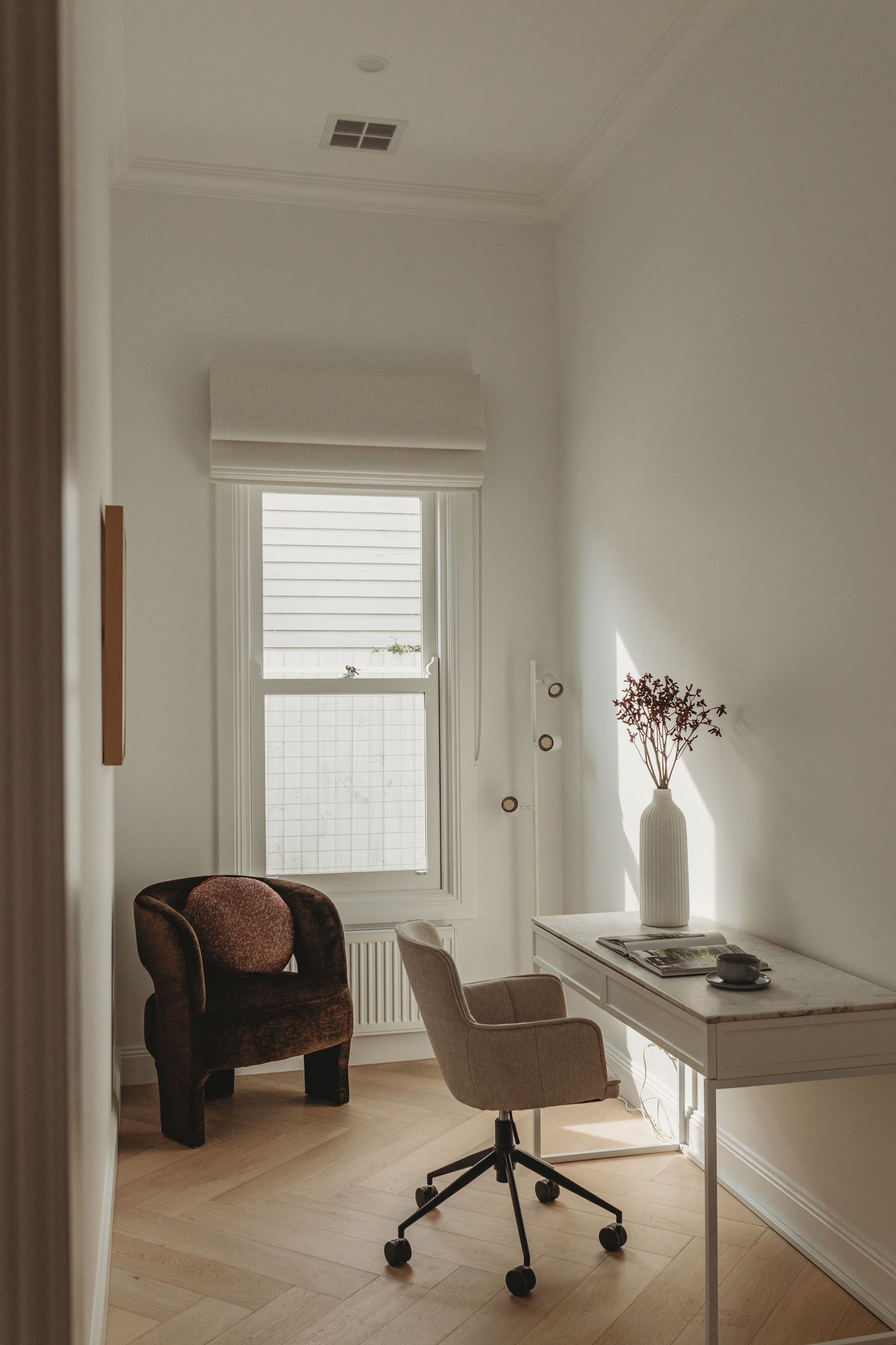 Minimalist home office with herringbone floors, a beige desk chair, white desk with dried floral vase, and brown armchair by window.