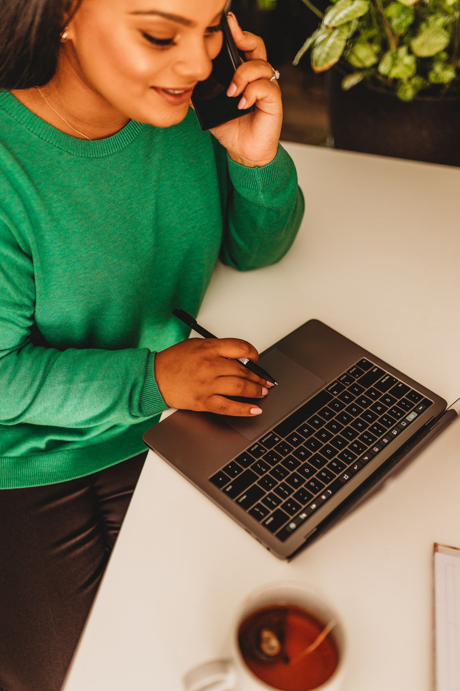 Woman in green sweater multitasking on laptop while talking on phone at a modern workspace.
