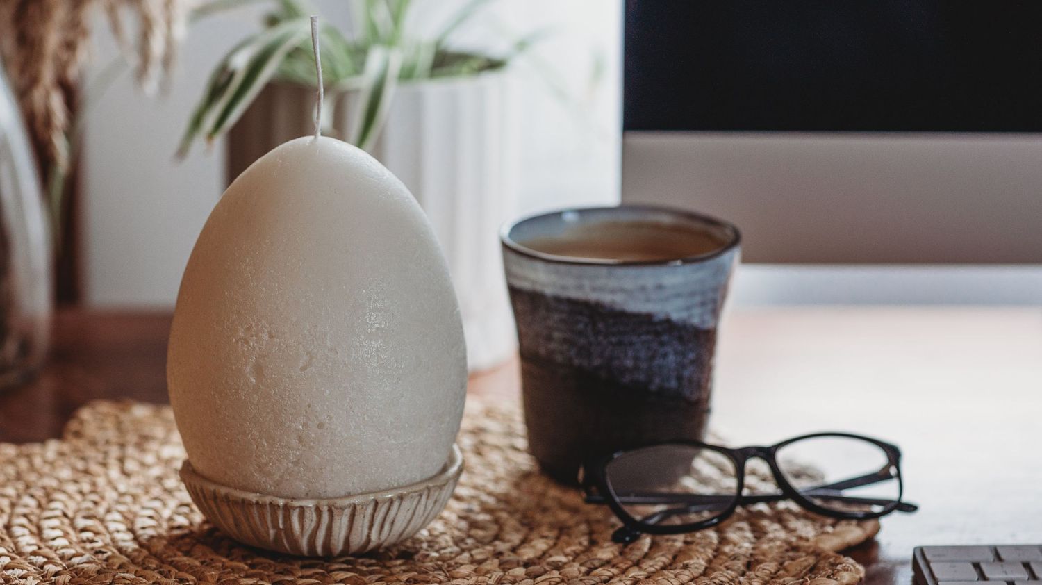 Egg-shaped white candle beside a ceramic coffee mug and glasses on a woven desk mat.