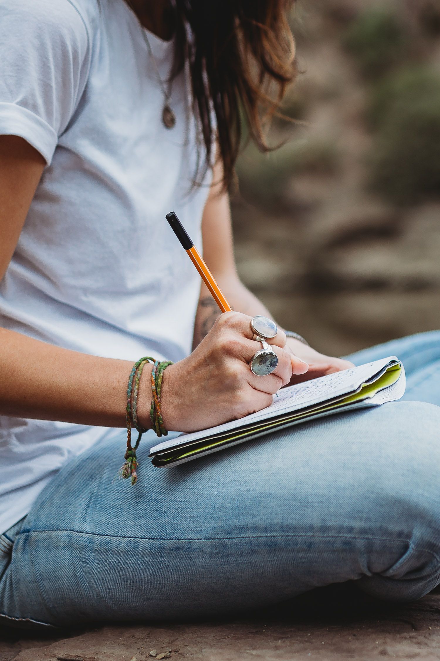 Woman in white tee and jeans writing in notebook outdoors wearing boho bracelets and rings.