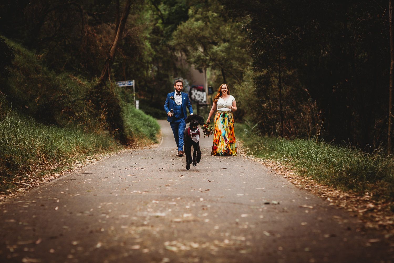 Joyful couple running with their black dog down a scenic tree-lined wedding venue path.