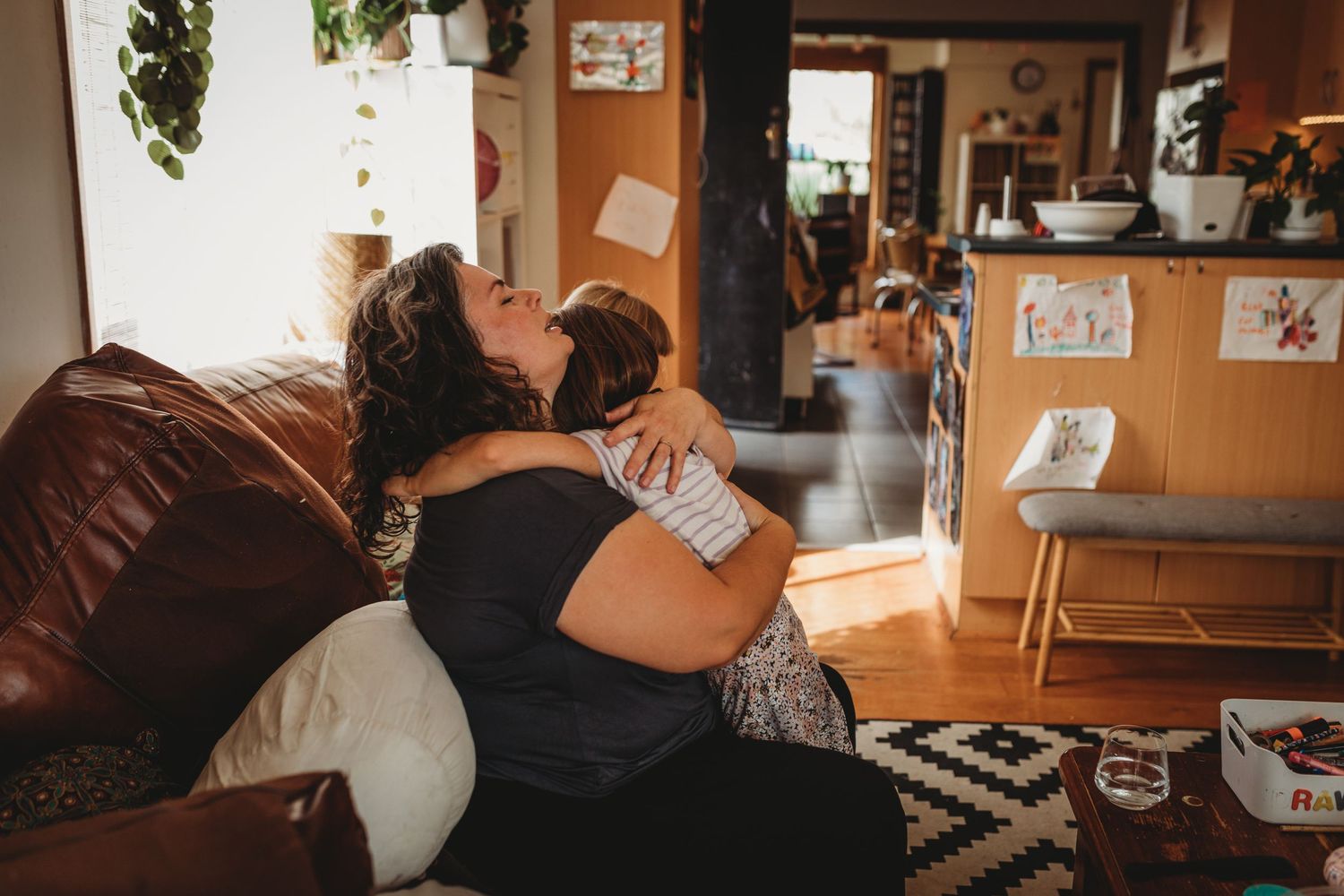 A mother sits on a living room floor hugging her young child on a cozy sunny afternoon.