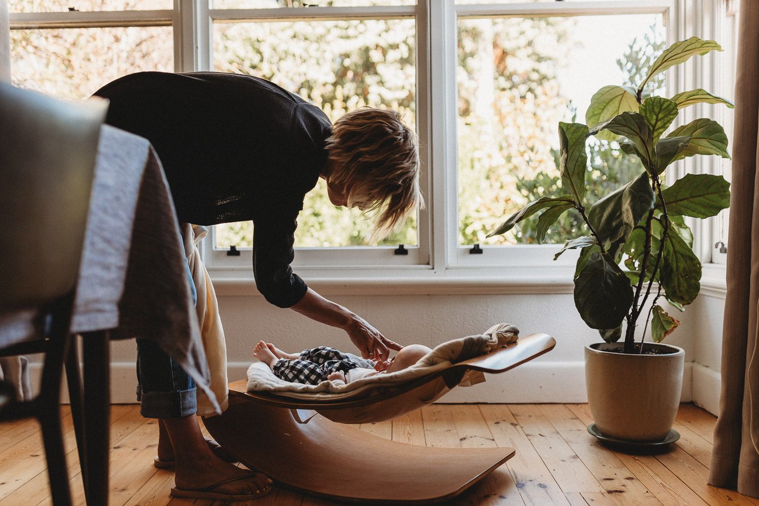Parent gently tending to newborn baby in wooden rocker chair near sunny window indoors.