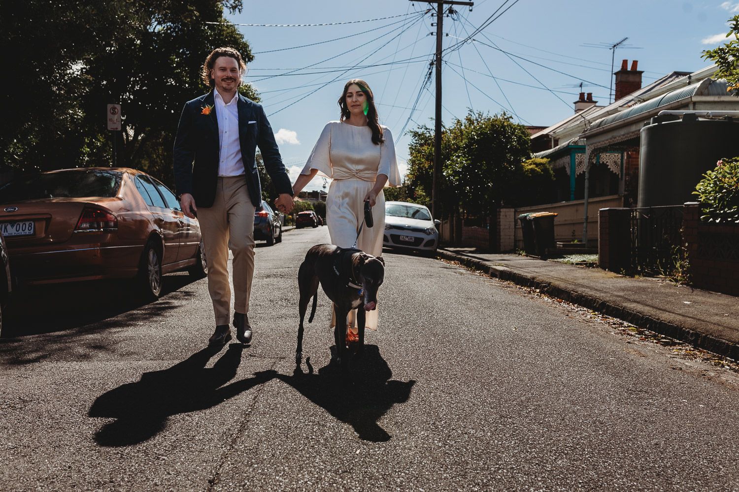 A couple holding hands walking their dog down a suburban street on a sunny day.