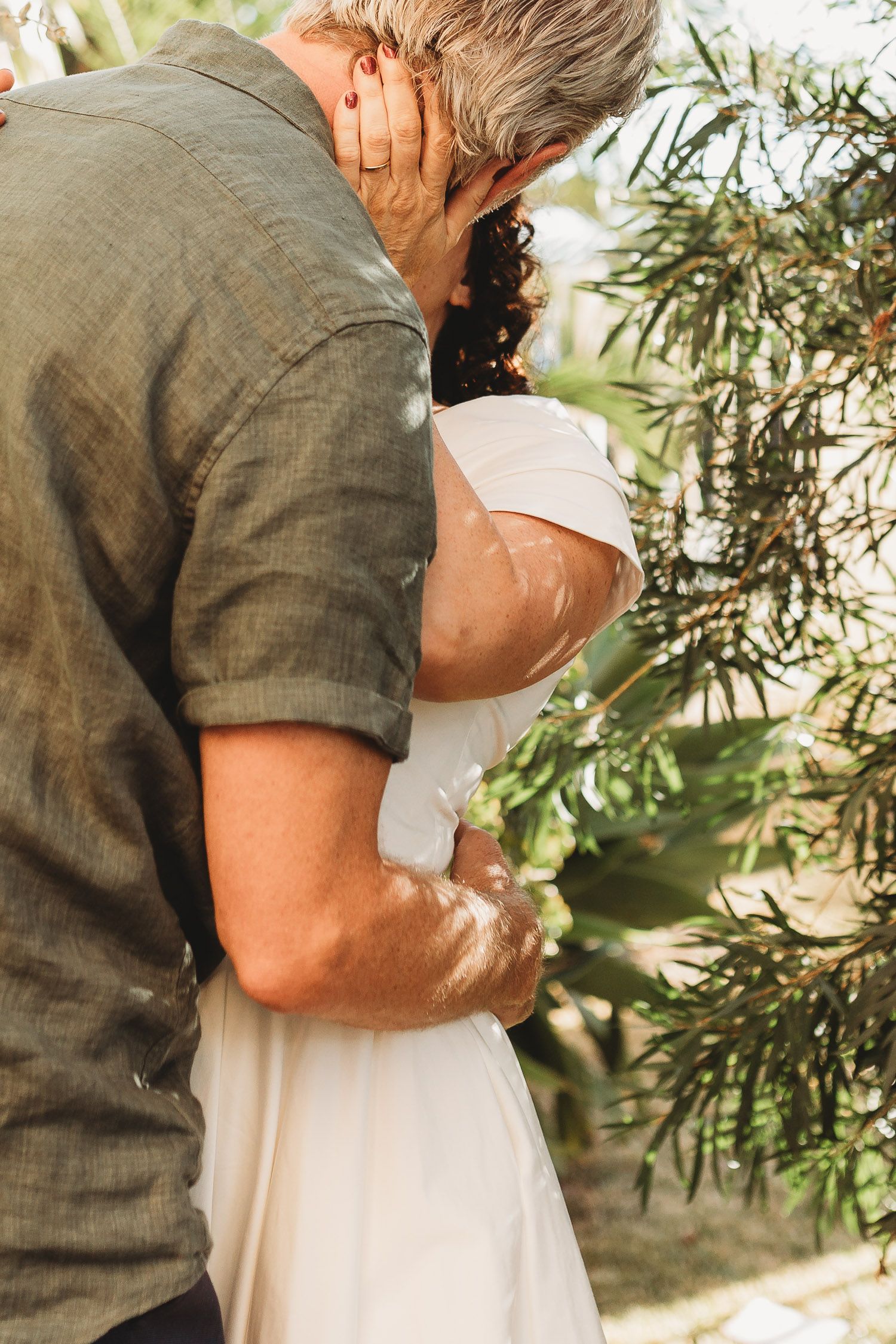 Groom in green shirt tenderly embracing bride in white dress among tropical garden greenery.