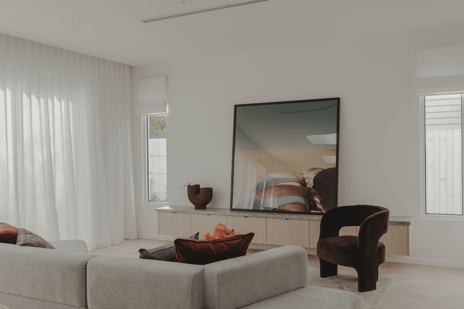 Bright minimalist living room with grey sofa, dark armchair, large artwork above console, and sheer white curtains.