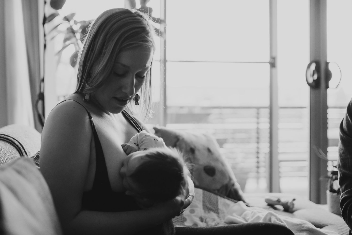 Black and white image of blonde mother breastfeeding newborn baby by bright window.