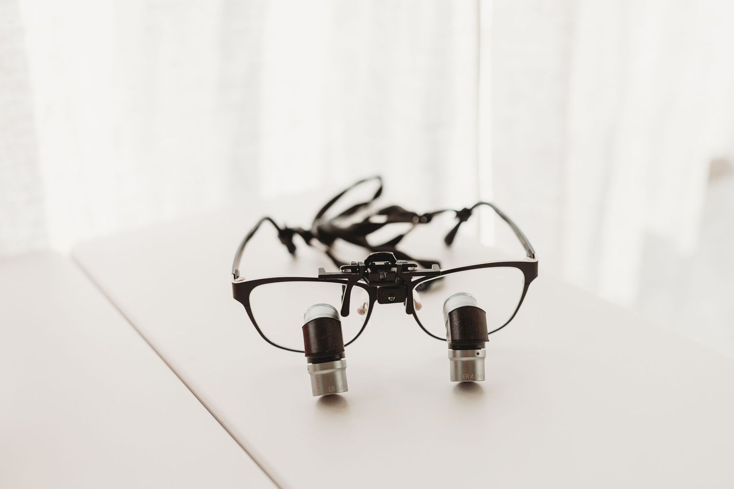 Dental loupes magnifying glasses sitting on white table near sheer curtain.
