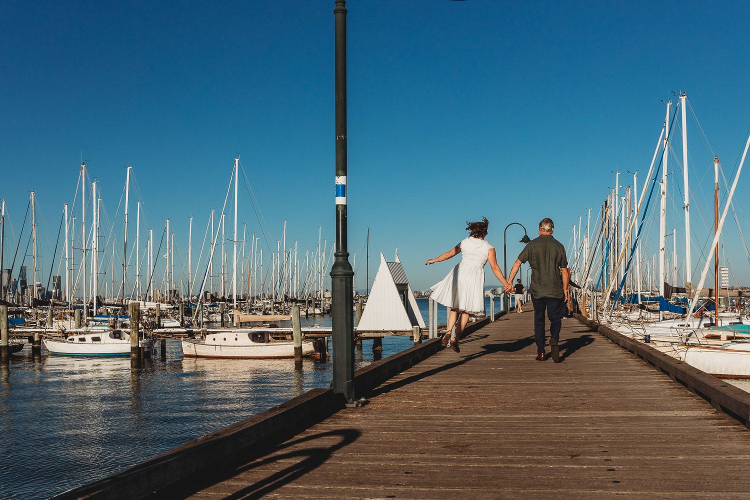 Couple walking hand in hand along a wooden marina pier surrounded by sailboats on a clear blue day.