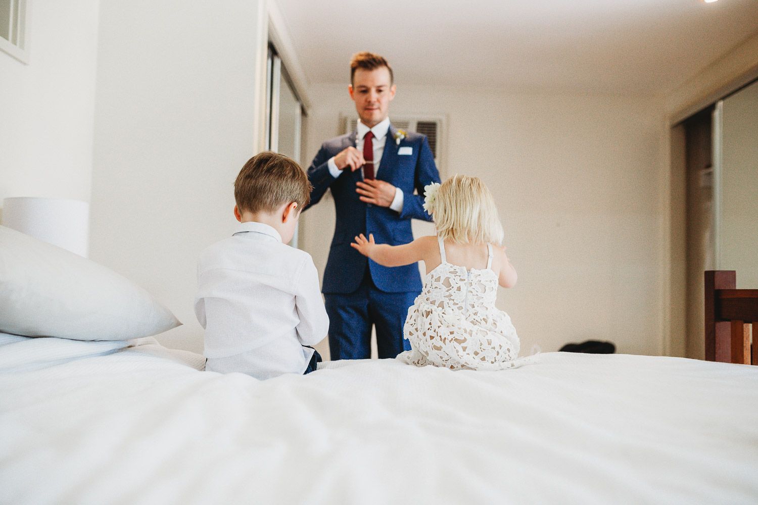 Groom in blue suit interacting with two young children on a white bed.