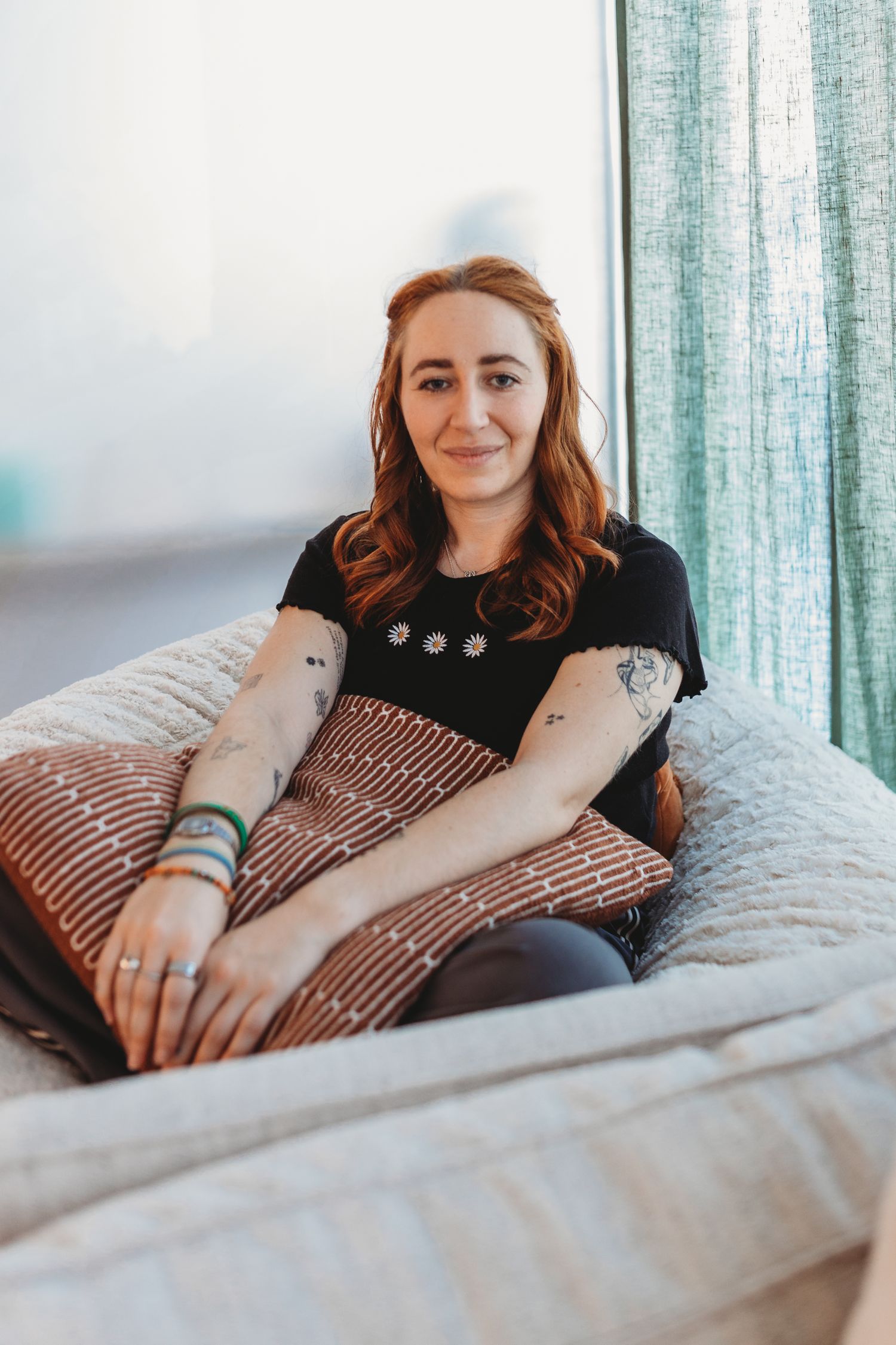Smiling redhead woman with tattoos sitting on a cosy bed with striped pillow near a teal curtained window.