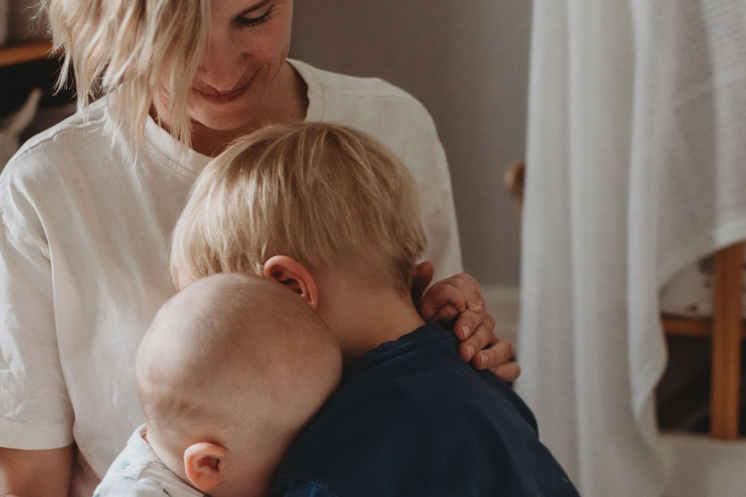 A mother smiles warmly while embracing two young blonde children in a cozy indoor setting.